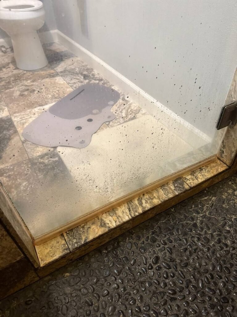 Shower glass panel with dark spots and haze and a stained threshold above a black pebble floor before JenyClean cleaning in Redmond, WA