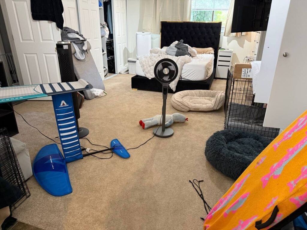 Carpeted bedroom with boards, a fan, dog beds and items on the floor before JenyClean monthly cleaning in Redmond, WA