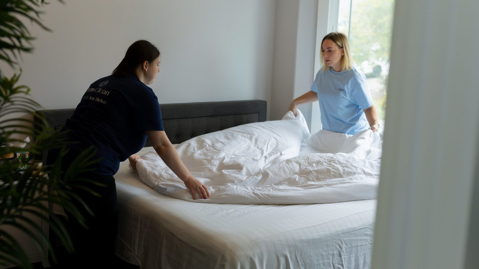 Two JenyClean cleaners changing white bed linen in a bedroom